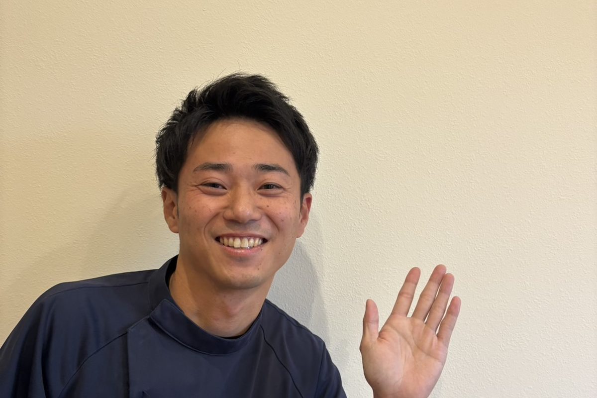 白い壁の前で、片手を挙げて笑顔であいさつする安井先生
