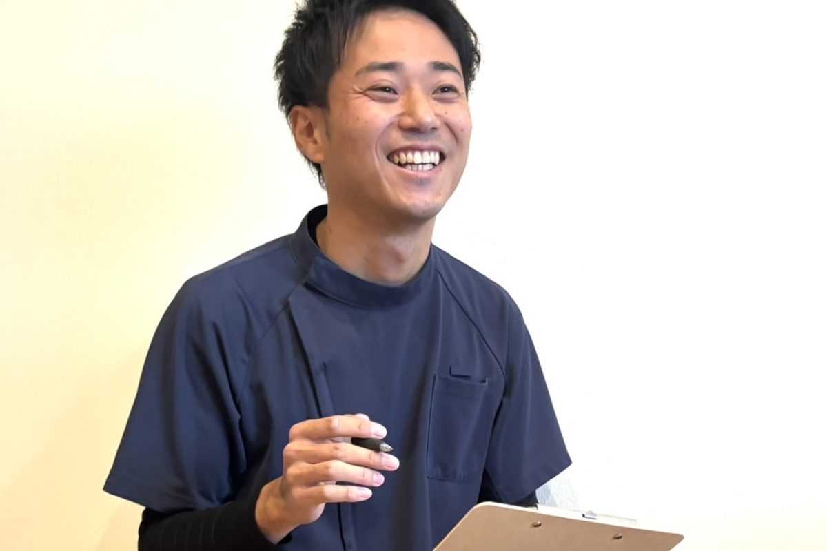 クリップボードとペンを手に、ひざをついて笑顔で話を聞く安井先生